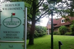 Willkommen-Schild für einen Golfclub mit einer roten Fahne, einer Laterne und einem Haus im Hintergrund.