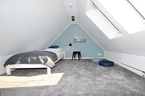 Ein Schlafzimmer mit schrägemausstem Dach, einem Bett mit grauer Decke, einem kleinen Hocker und einem runden, blauen Gegenstand auf dem Boden.