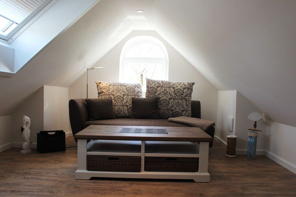 A cozy attic bedroom with a sloped ceiling, a wooden bed frame, a large window, and a small table with storage drawers.