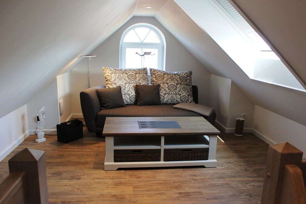 Ein gemütliches Schlafzimmer im Loft-Stil mit schrägem Dach, einem großen Fenster, einer Couch mit gemusterten Kissen, einem Holztisch und einem kleinen Beistelltisch.