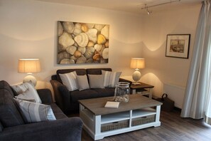 A living room with a sofa, a coffee table, and two lamps. There is a painting on the wall and a framed picture above the lamp. - Holiday home with 2 bedrooms (Greetsiel)
