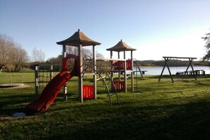 Ein Spielplatz mit Rutsche und Klettergerüsten, einem Picknicktisch und Blick auf ein Gewässer.