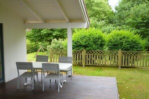 Eine überdachte Terrasse mit Tisch und Stühlen, umgeben von einem Holenzaun und üppiger Bepflanzung.
