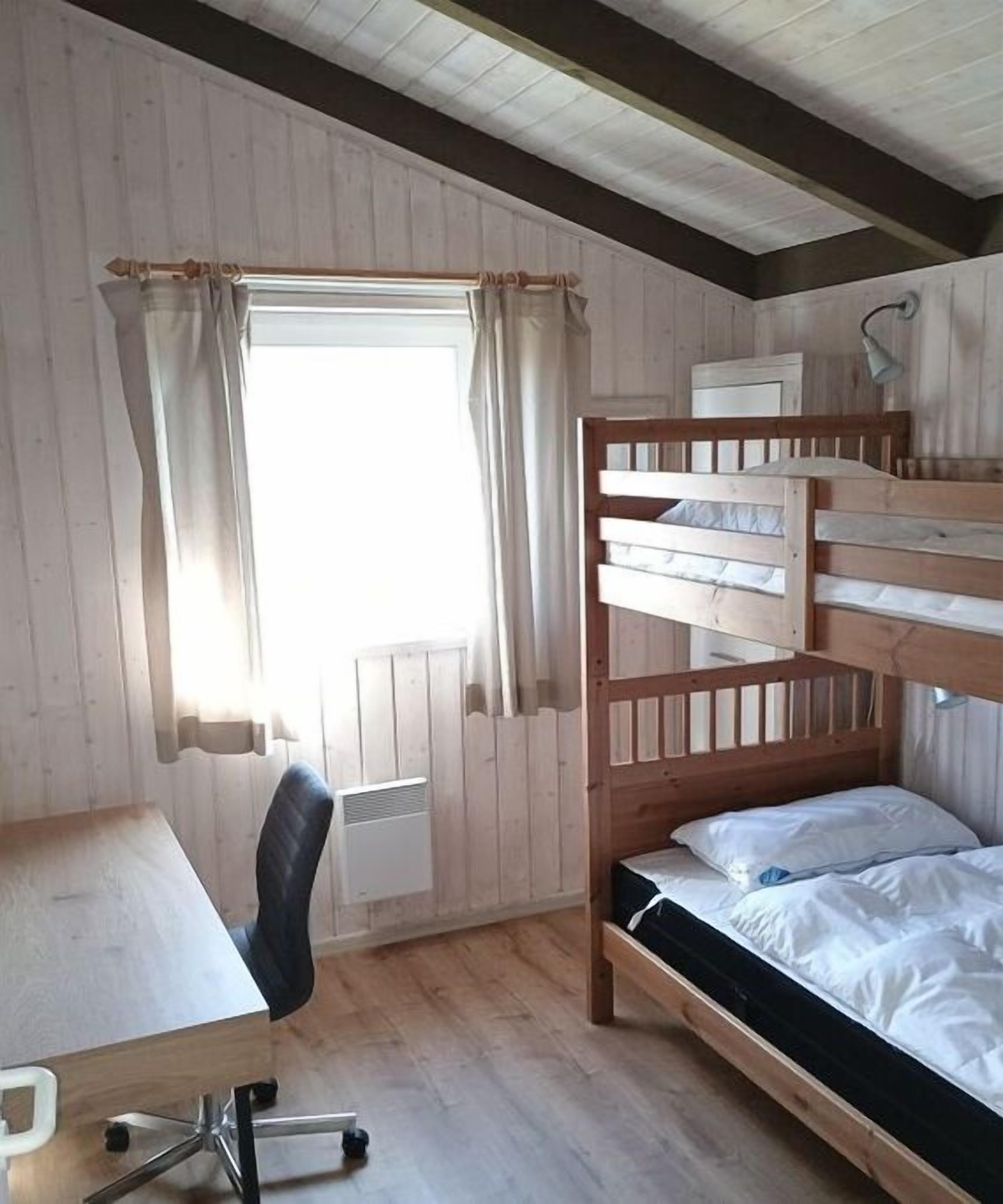 Une chambre avec un lit superposé, un bureau avec une chaise, une fenêtre avec des rideaux et un radiateur.
