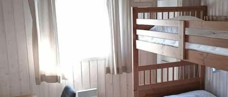 A room with a bunk bed, a desk with a chair, a window with curtains, and a radiator.