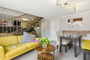 A modern living room with a yellow sofa, a round coffee table with a vase, a dining area with a table and chairs, and a staircase with a metal railing. - Holiday home with 1 bedroom (Wieck\/Darß)