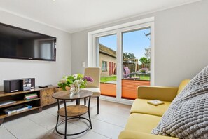 Ein modernes Wohnzimmer mit Flachbildfernseher, einem runden Tisch mit Blumenvase, einem gelben Sofa und einer Glastür zum Außenbereich.