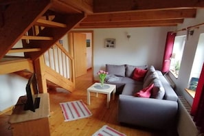 A living room with a grey sofa, a small white table with a vase of flowers, and a wooden staircase. - Holiday home with 2 bedrooms (Krautsand)