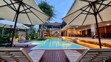 A pool area with wooden loungers and umbrellas, surrounded by a landscaped garden and a building with a balcony.