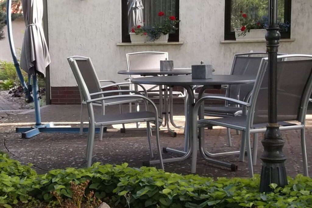 Ein Sitzbereich im Freien mit Metallstühlen und einem Tisch, umgeben von Pflanzen und einem Gebäude mit Fenstern.
