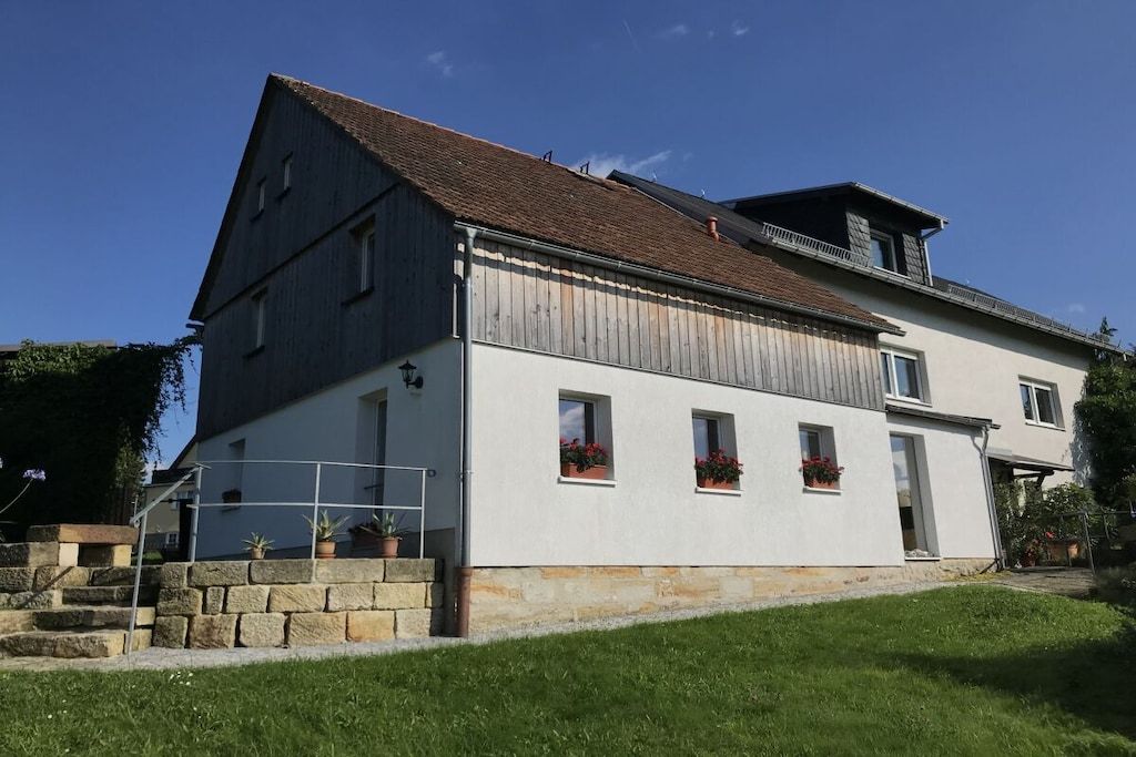 A two-story house with a sloped roof, a balcony on the upper floor, and a stone staircase leading to the entrance.