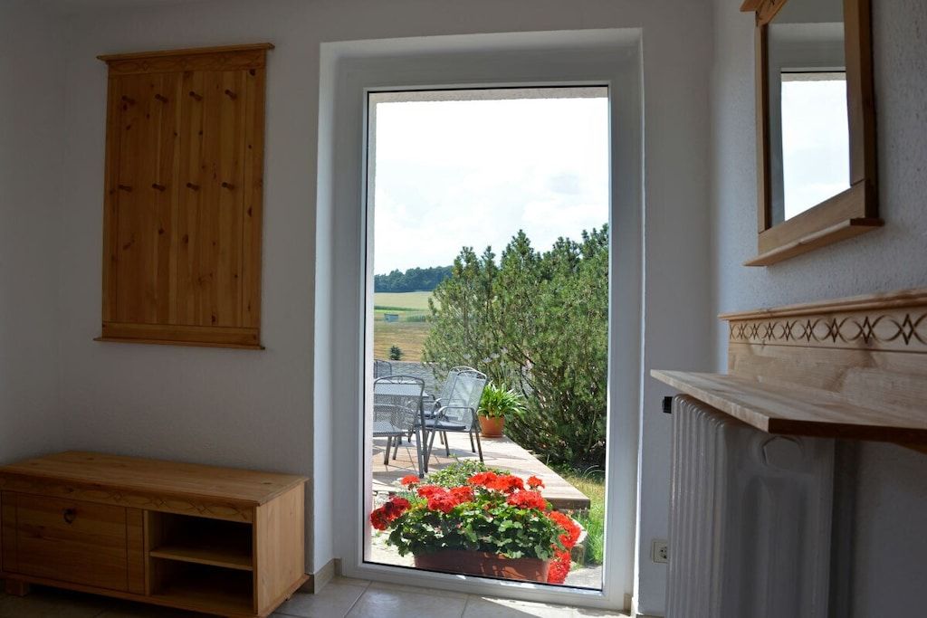 A room with a wooden cabinet, a bench, and a view through a glass door to an outdoor patio with chairs and a flower pot.