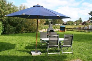 Un espace de restauration en plein air avec une table et des chaises sous un grand parasol, un four en pierre et un décor rustique agrémenté d’arbres et d’un bâtiment en arrière-plan.