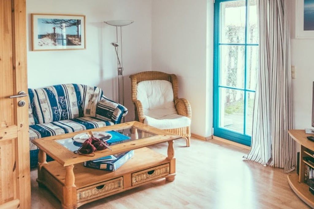 Ein gemütliches Wohnzimmer mit einem gestreiften Sofa, einem Korbstuhl, einem Holztisch und einem großen Fenster mit Vorhängen.