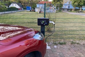 Une voiture rouge est garée devant une clôture, sur un trottoir pavé de briques, à proximité d’une borne de recharge portant l’inscription « E3 DC ».