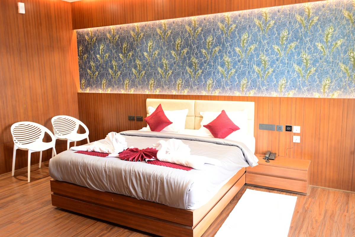 Habitación de hotel con una cama grande, dos sillas blancas, un cabecero de madera y una pared decorada sobre la cama.
