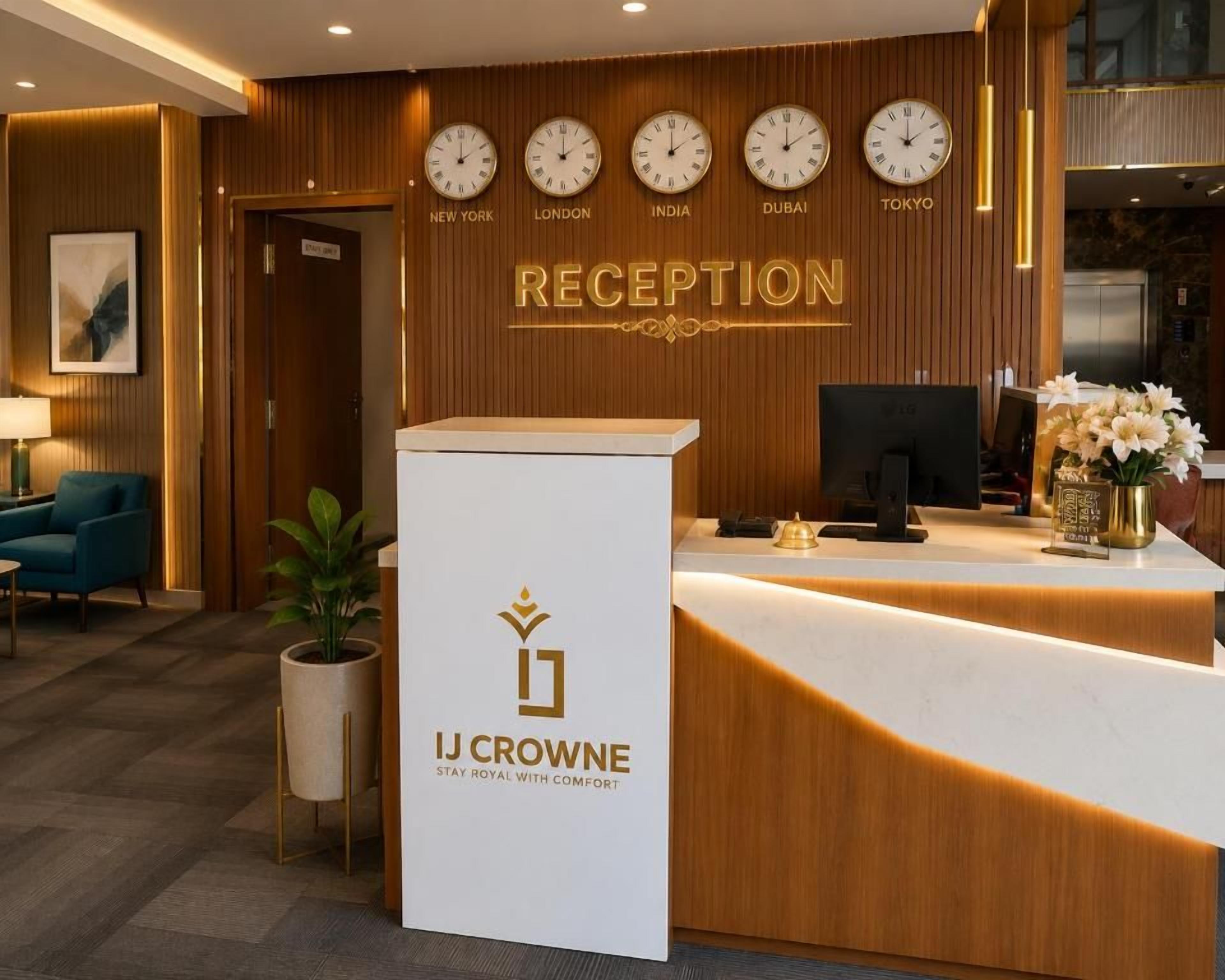 Uma área de recepção com balcão, relógios mostrando a hora em Nova York, Londres, Índia, Dubai e Tóquio, e um letreiro com a inscrição 'IJ CROWNE STAY ROYAL WITH COMFORT'.