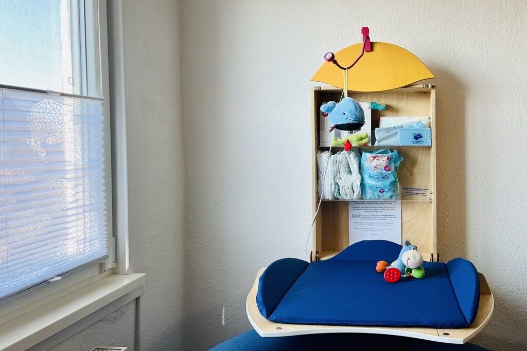 Una habitación con ventana, una cama azul con colchón azul, un armario de madera con espacio de almacenamiento y un estante pequeño con juguetes.