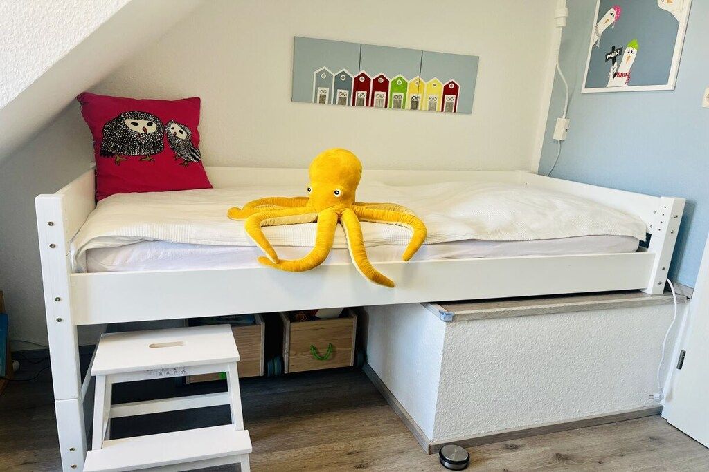 Un lit superposé avec un oreiller rouge, un jouet en forme de poulpe jaune et une décoration murale représentant des maisons colorées.