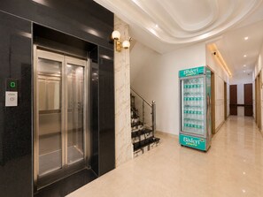 Un ascensor moderno con pantalla digital, una escalera con barandilla y una máquina expendedora que muestra el logo de una botella de agua.