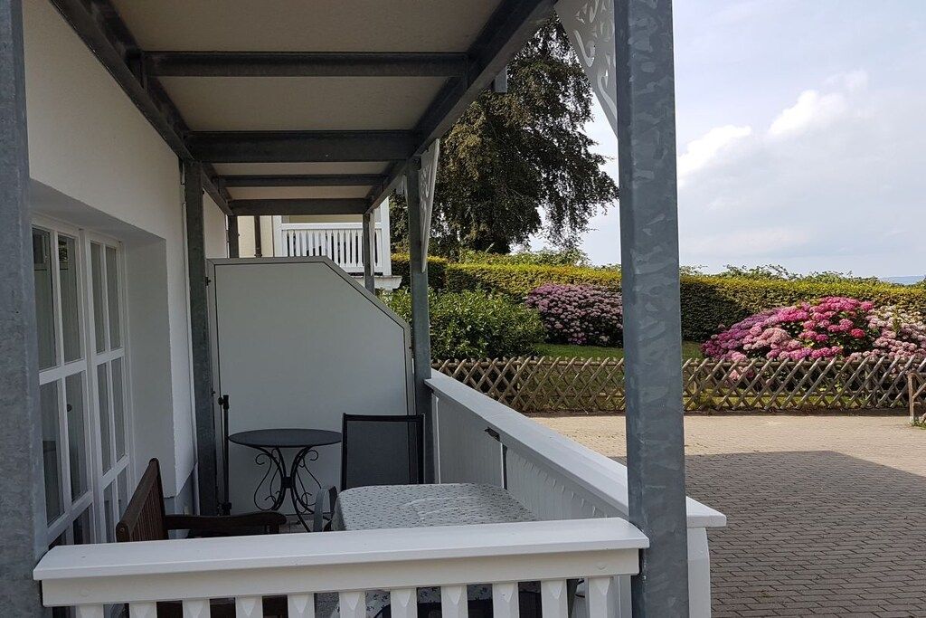 A covered outdoor area with a white railing, a table, and chairs, leading to a building entrance.