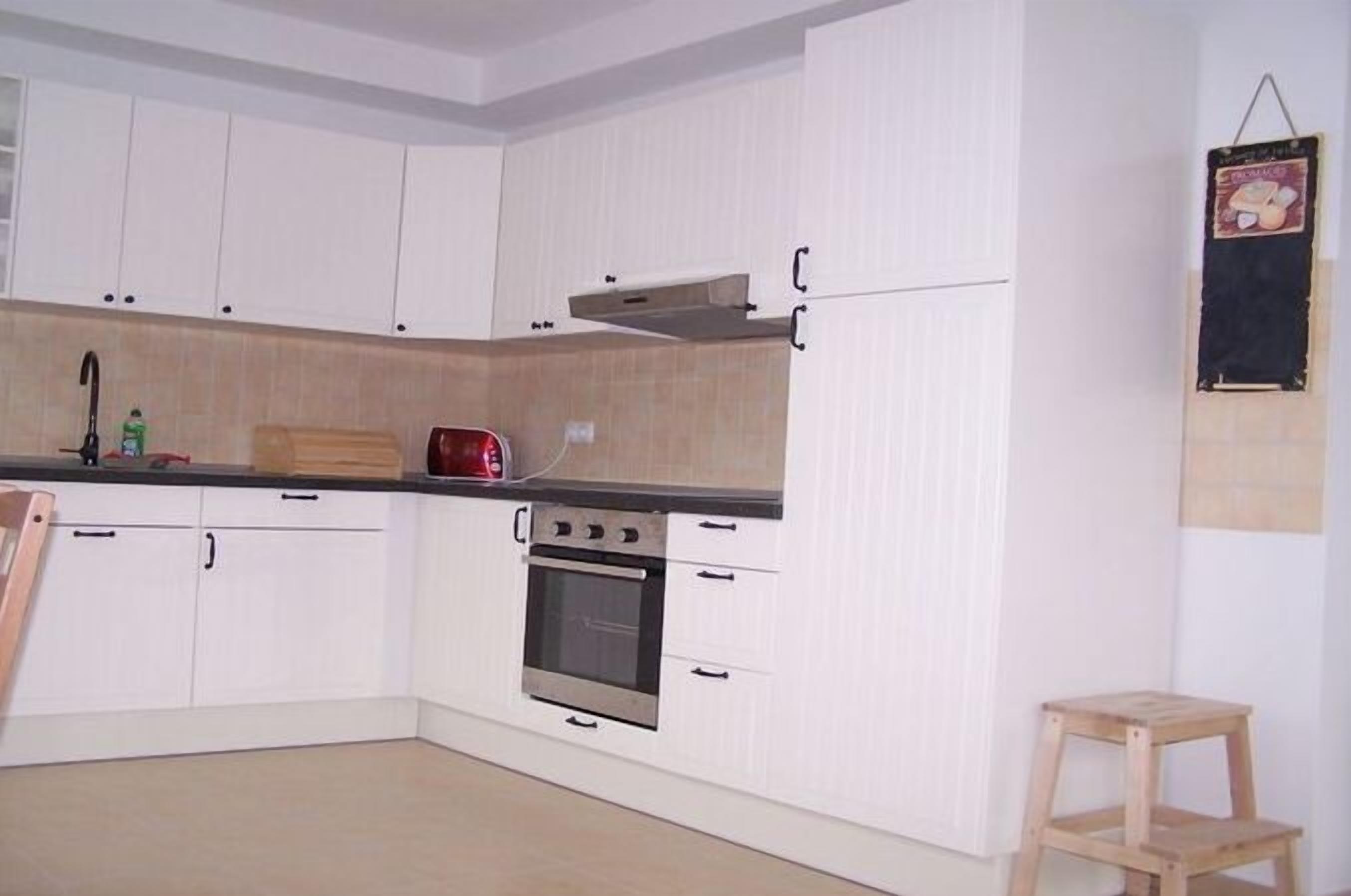 Une cuisine avec des meubles blancs, un four encastré, un grille-pain et un tabouret en bois.