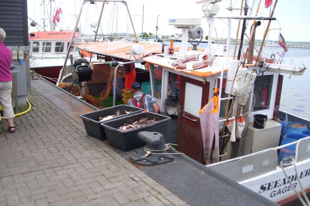 A boat docked at a marina with fishing equipment, including two large black containers with fish, a winch, and a person standing by.