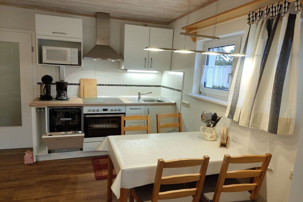 A compact kitchen with a microwave, stove, and sink, wooden chairs, and a table with a white tablecloth.