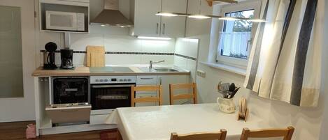 A compact kitchen with a microwave, stove, and sink, wooden chairs, and a table with a white tablecloth.