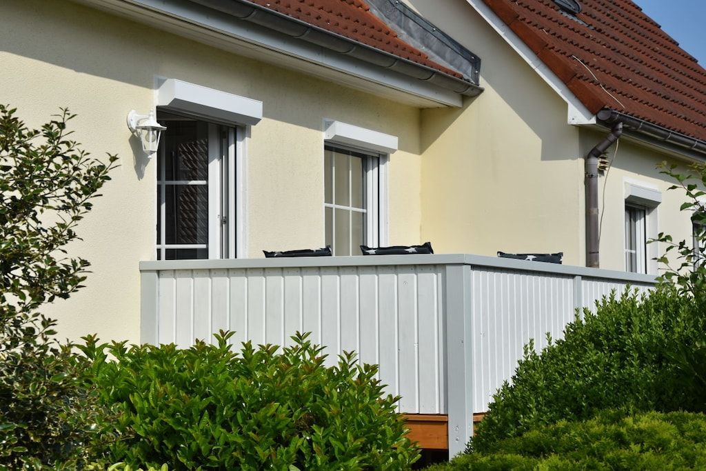 Une maison à l’extérieur blanc, avec un toit recouvert de tuiles rouges et une véranda blanche dotée d’une balustrade.