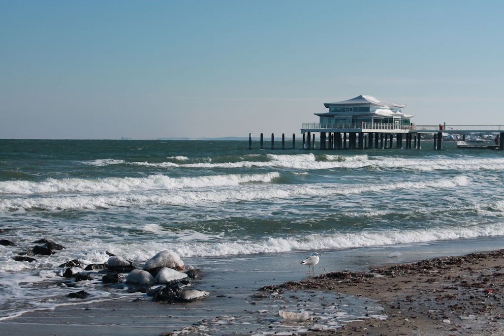 A coastal building with a pier extending into the sea, surrounded by waves and rocks.