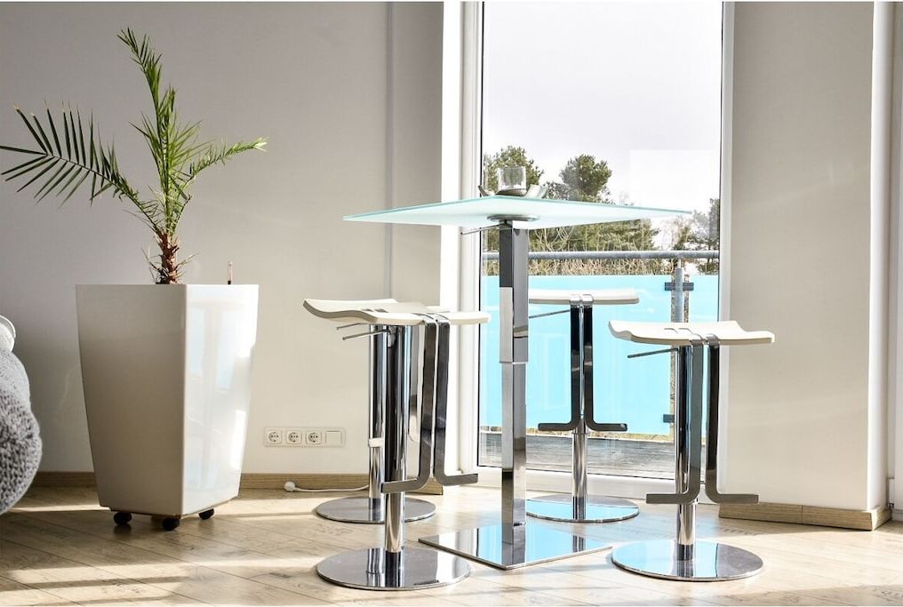 A modern room with a glass-top table and two bar stools, a potted plant, and a view of a balcony with a pool.