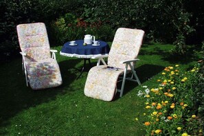 Two chairs with floral upholstery, a round table with a blue tablecloth, and a garden with flowers and greenery. - Holiday apartment with 2 bedrooms (Borgsum auf Föhr)