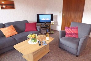 A living room with a sofa, armchair, wooden coffee table, television, remote control, wine bottle, and a bouquet of flowers. - Holiday apartment with 2 bedrooms (Borgsum auf Föhr)