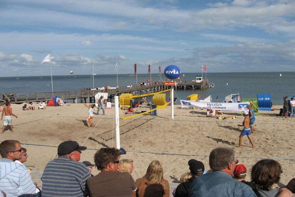 Ein Beachvolleyballspiel mit Zuschauern, einem Steg und einem Rettungsturm im Hintergrund.