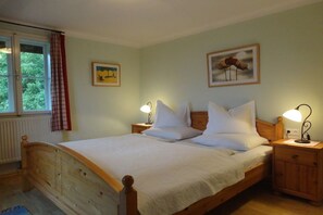 A single bed with a wooden headboard and footboard, flanked by two bedside tables with lamps. A window with a view of greenery is visible, and two framed pictures adorn the wall. - Landhaus Arztmann - Fewo Linde (Bodensdorf am Ossiacher See)