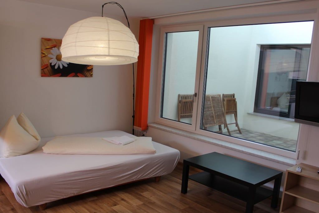 A bedroom with a bed, a bedside table, a window with a view of outdoor furniture, and a ceiling light fixture.