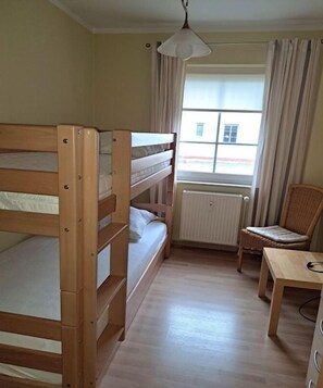 A room with bunk beds, a chair, a table, a window with curtains, and a radiator. - House on the Reef || Ostseebad Prerow (Ostseebad Prerow)