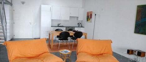 A modern living room with orange sofas, a dining area, and a kitchen in the background.