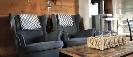 Une pièce comprenant deux fauteuils gris, une table basse en bois agrémentée d’un élément décoratif, une cheminée et une étagère avec divers objets.