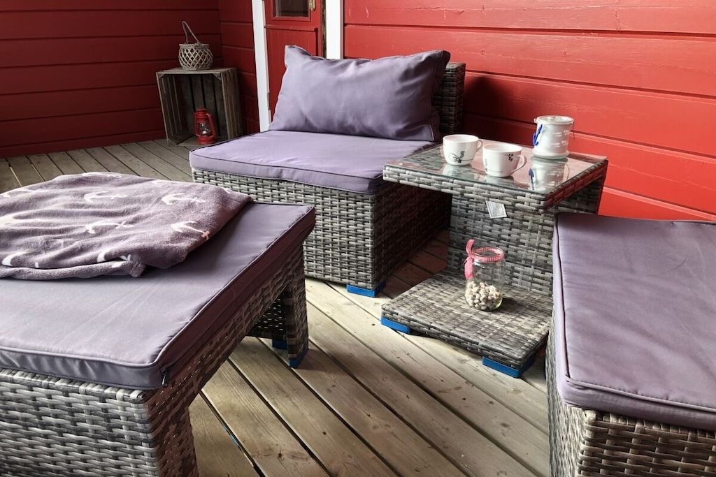 A cozy outdoor seating area with wicker furniture and cushions, a small table with cups and a decorative item, and a red wall in the background.