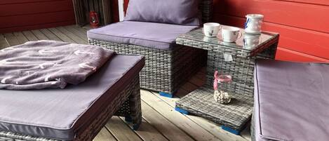 A cozy outdoor seating area with wicker furniture and cushions, a small table with cups and a decorative item, and a red wall in the background.