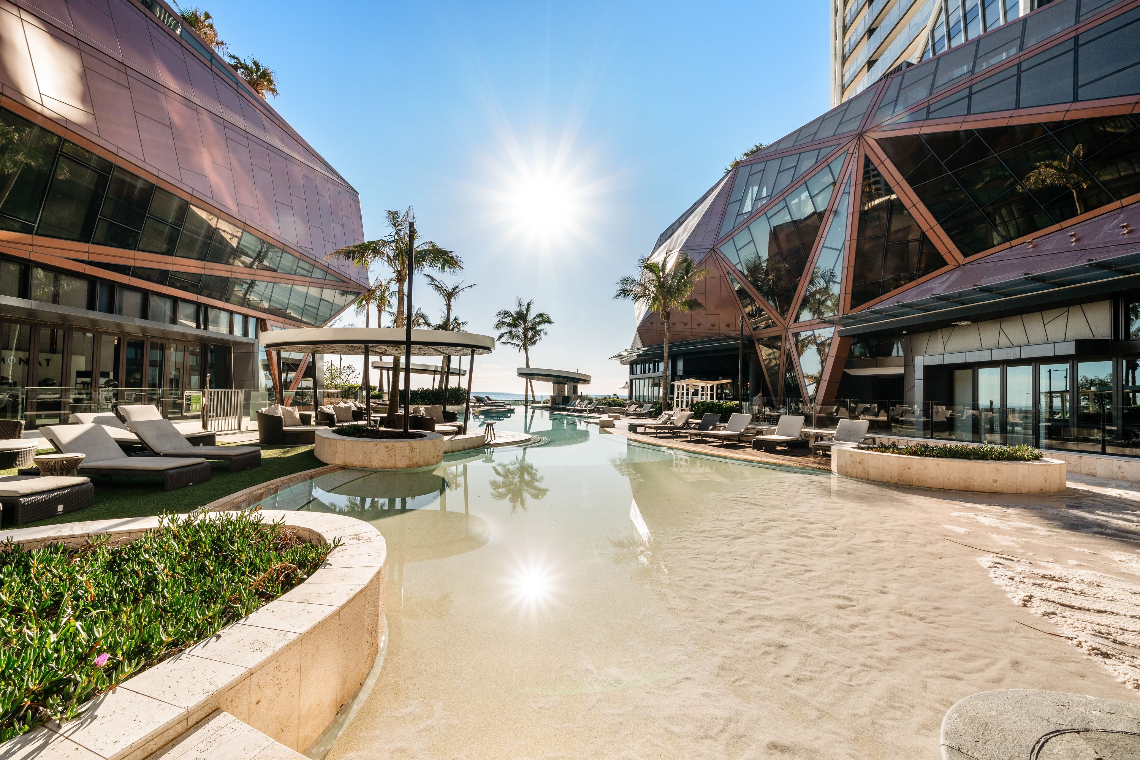 A modern hotel with a pool area, lounge chairs, and palm trees.