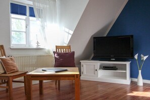 A living room with a wooden table, a television on a white stand, a window with blinds, and a vase with flowers. - Holiday home in Zingst with swimming pool (Zingst)
