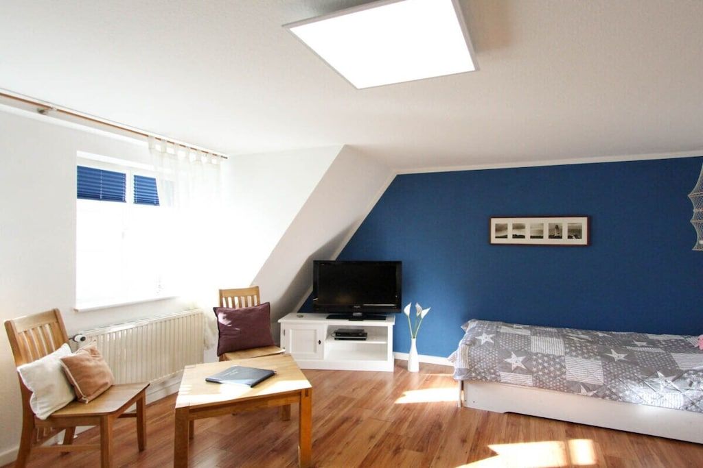 A room with a sloped ceiling, a television on a white cabinet, a sofa, a wooden table, and a window with blinds.