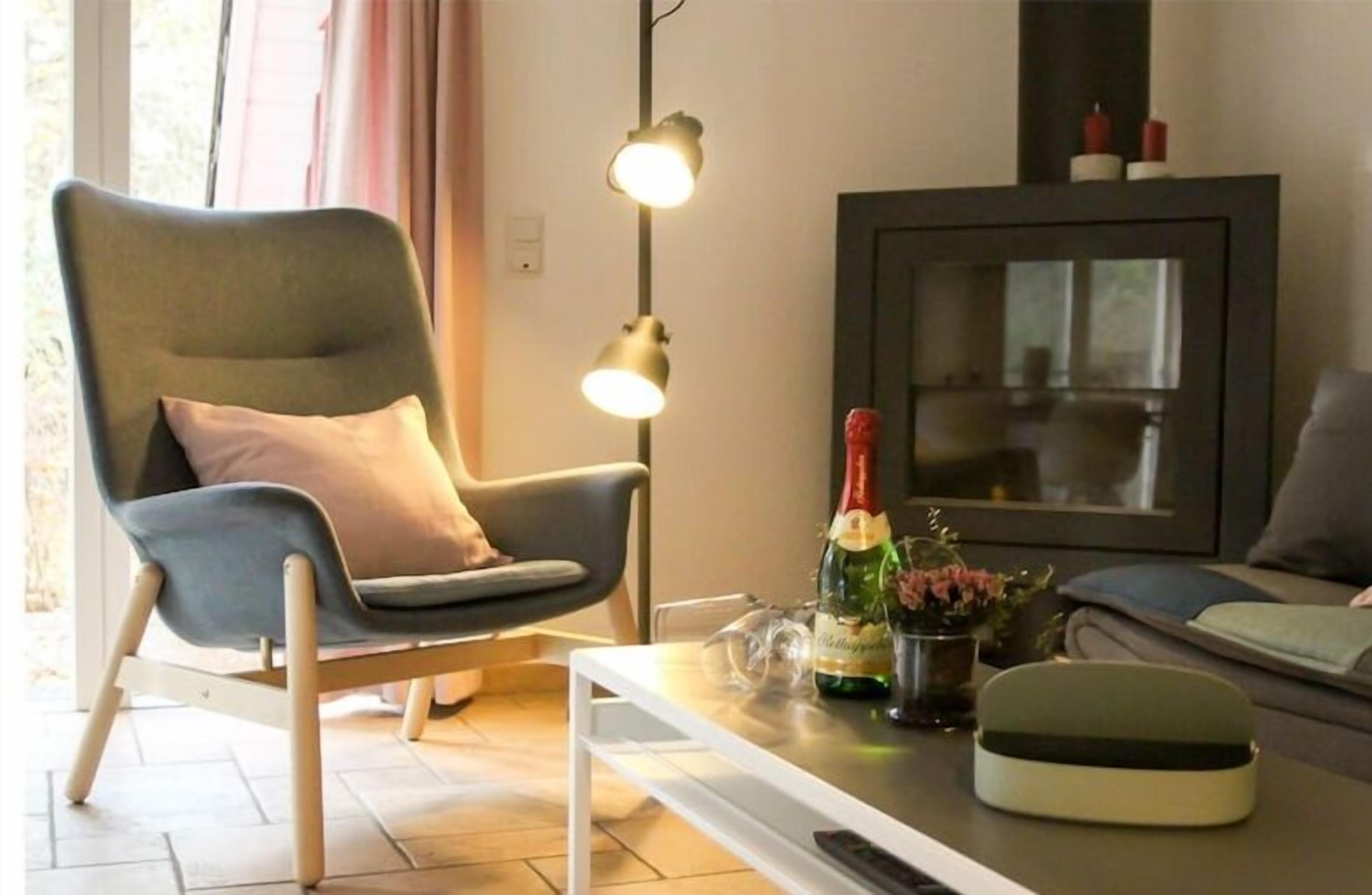 A modern living room with a comfortable armchair, a small table with a bottle and glasses, a television, and a framed picture on the wall.