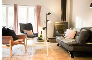 Ein Wohnzimmer mit Sofa, Sessel und einem kleinen Tisch mit einer Flasche. Eine Stehlampe und ein Kamin sind ebenfalls vorhanden.