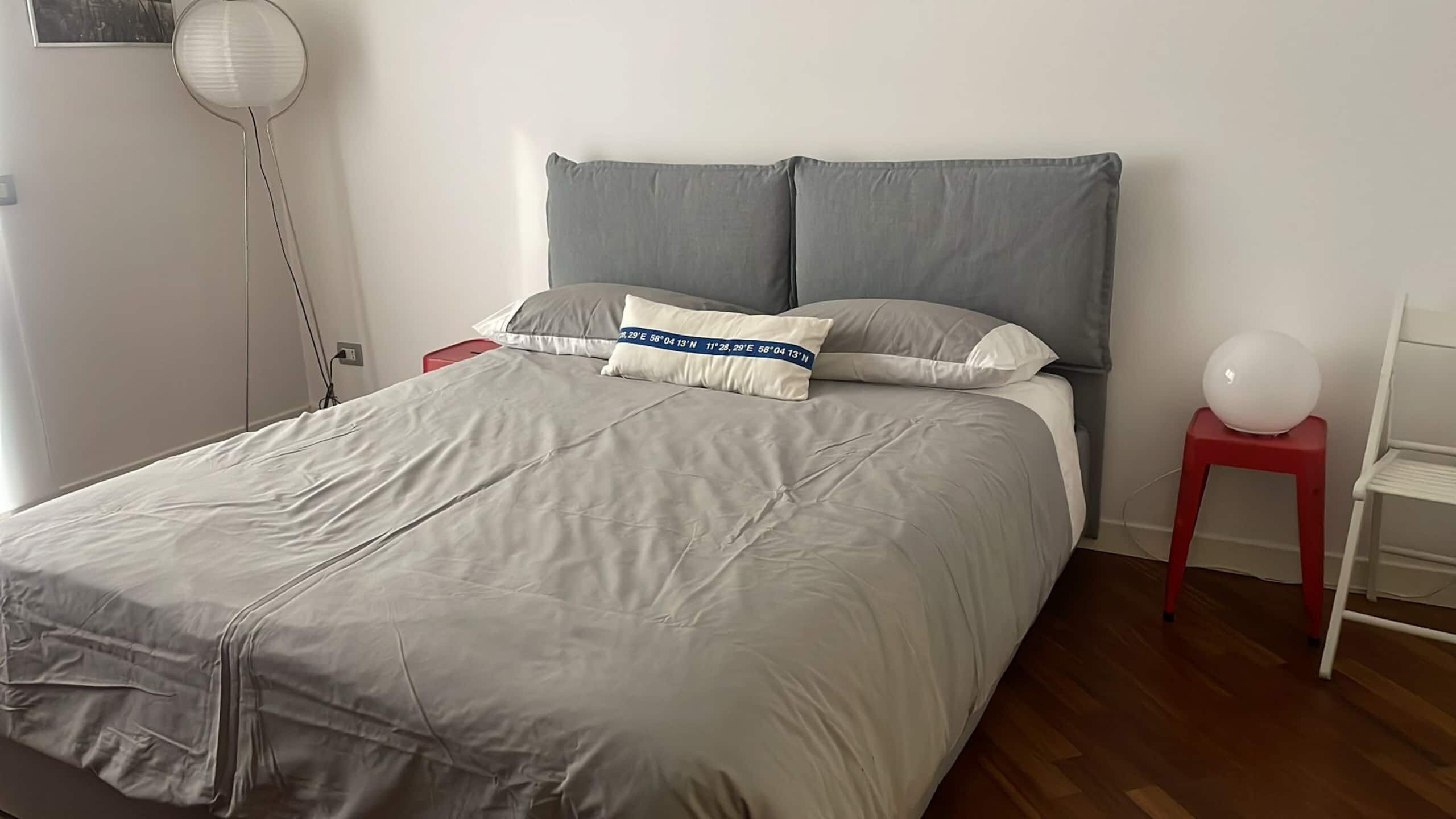Una cama bien hecha con edredón gris, una mesita de noche con un taburete rojo, y un objeto esférico blanco sobre el taburete.