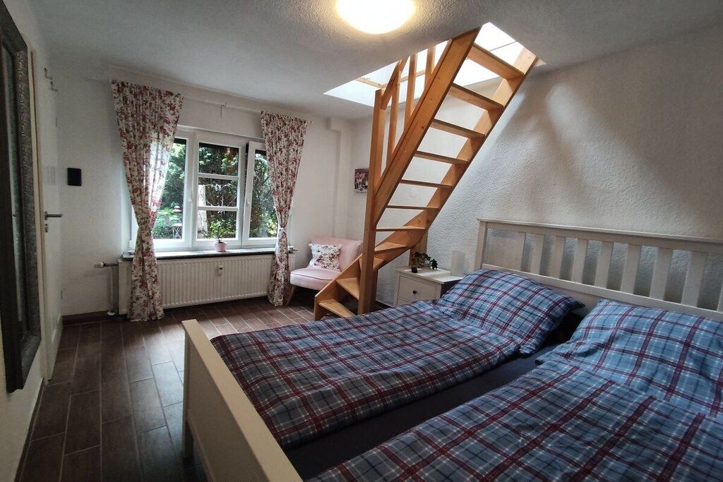 Et soveværelse med seng, et vindue med gardiner, en radiator, en trætrappe og et natbord.