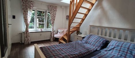 Et soveværelse med seng, et vindue med gardiner, en radiator, en trætrappe og et natbord.